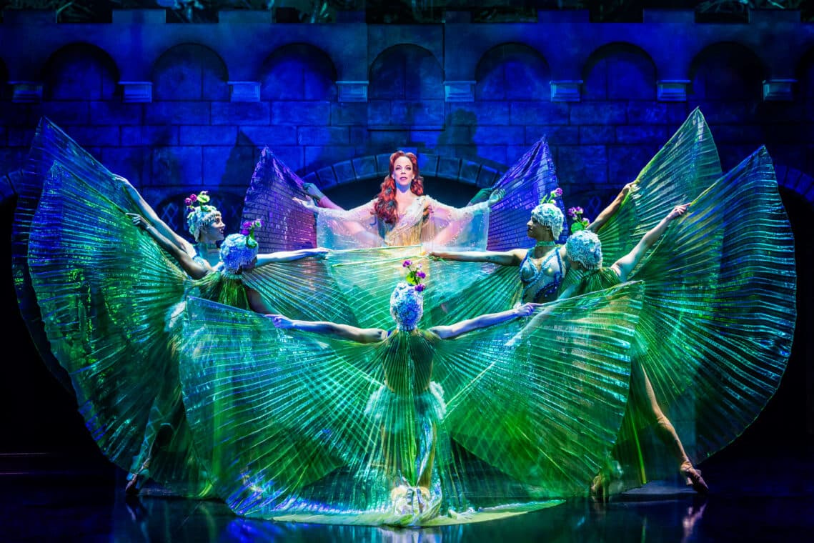 A woman in a white costume dancing with dancers in green costumes in the theater show titled 'Spamalot'