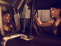 A woman driving a car, acting inside the Metropolitan Opera House, with her reflection visible in the side mirror