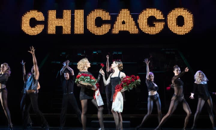 Two women holding roses, surrounded by several people, in the show of Chicago on Broadway at the Ambassador Theatre