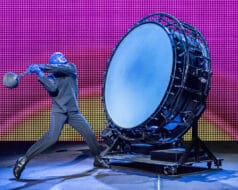 A man wearing a mask about to strike a very large bass drum in the Blue Man Group performance