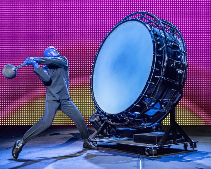 A man wearing a mask about to strike a very large bass drum in the Blue Man Group performance