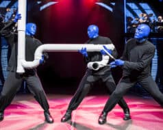 Three people in blue face paint are lifting a large pipe in the Blue Man Group performance