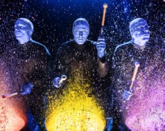 Three people wearing blue masks are playing musical instruments in the Blue Man Group performance