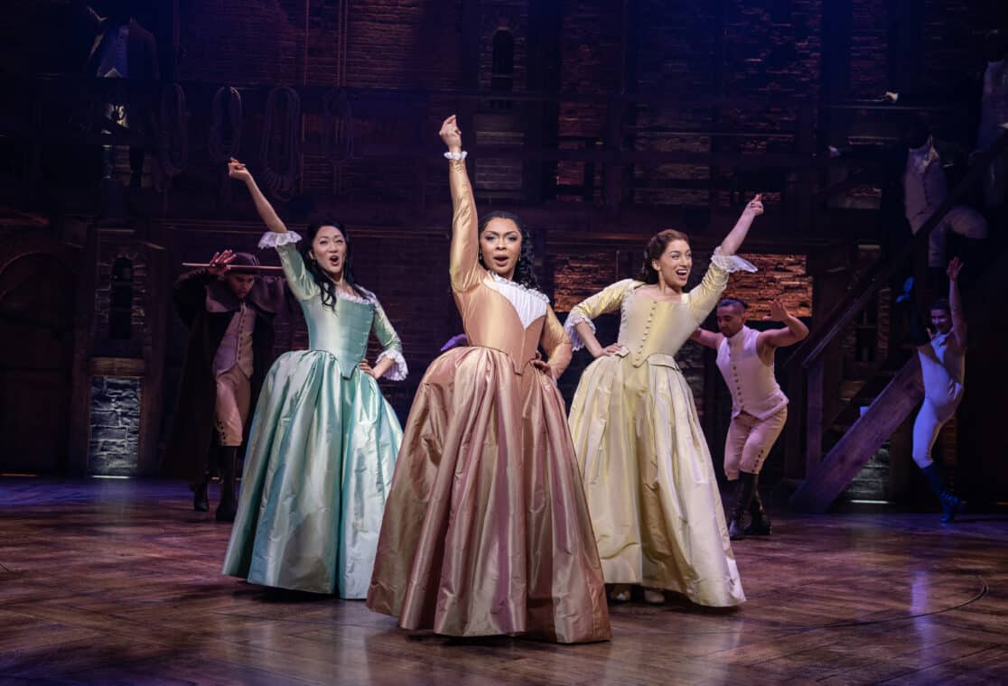Three women in colorful dresses dance with two men in the Broadway musical 'Hamilton'