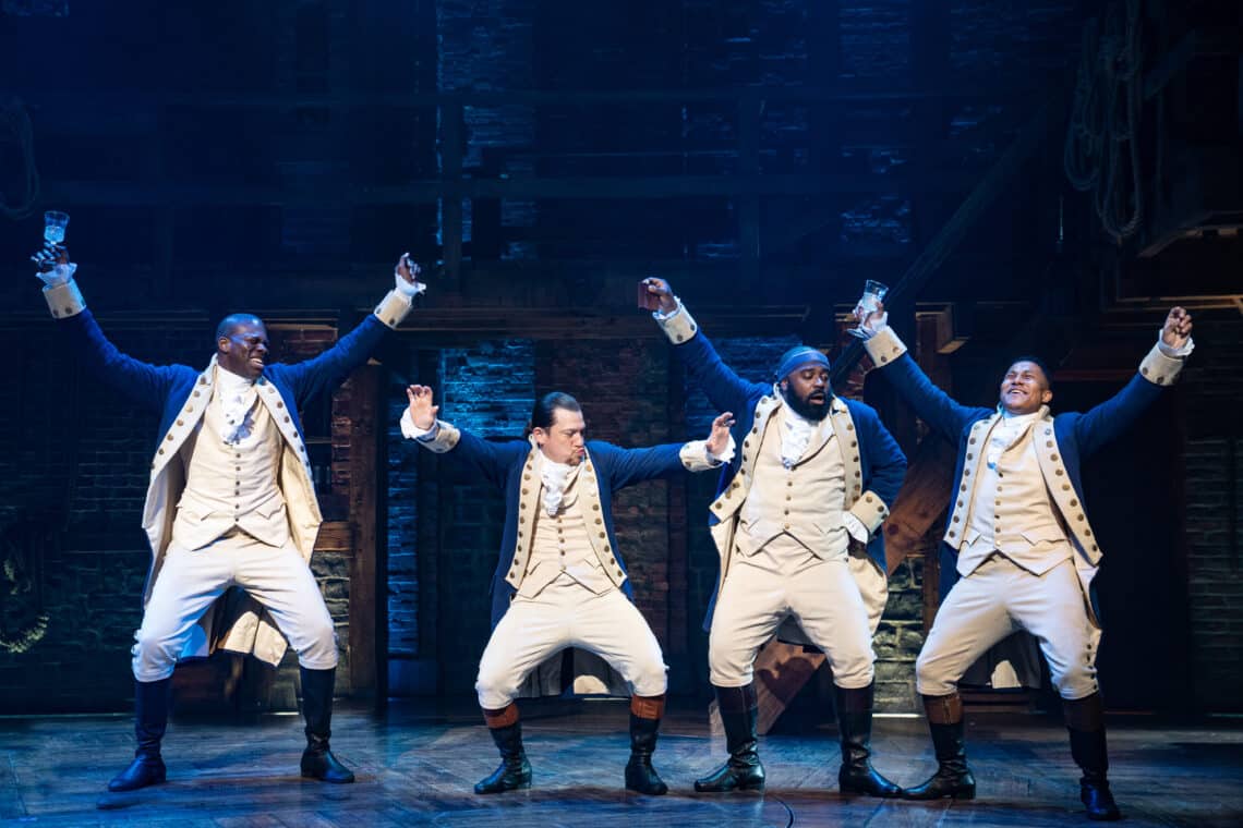 Four actors in blue costumes are dancing together in the Broadway musical titled Hamilton