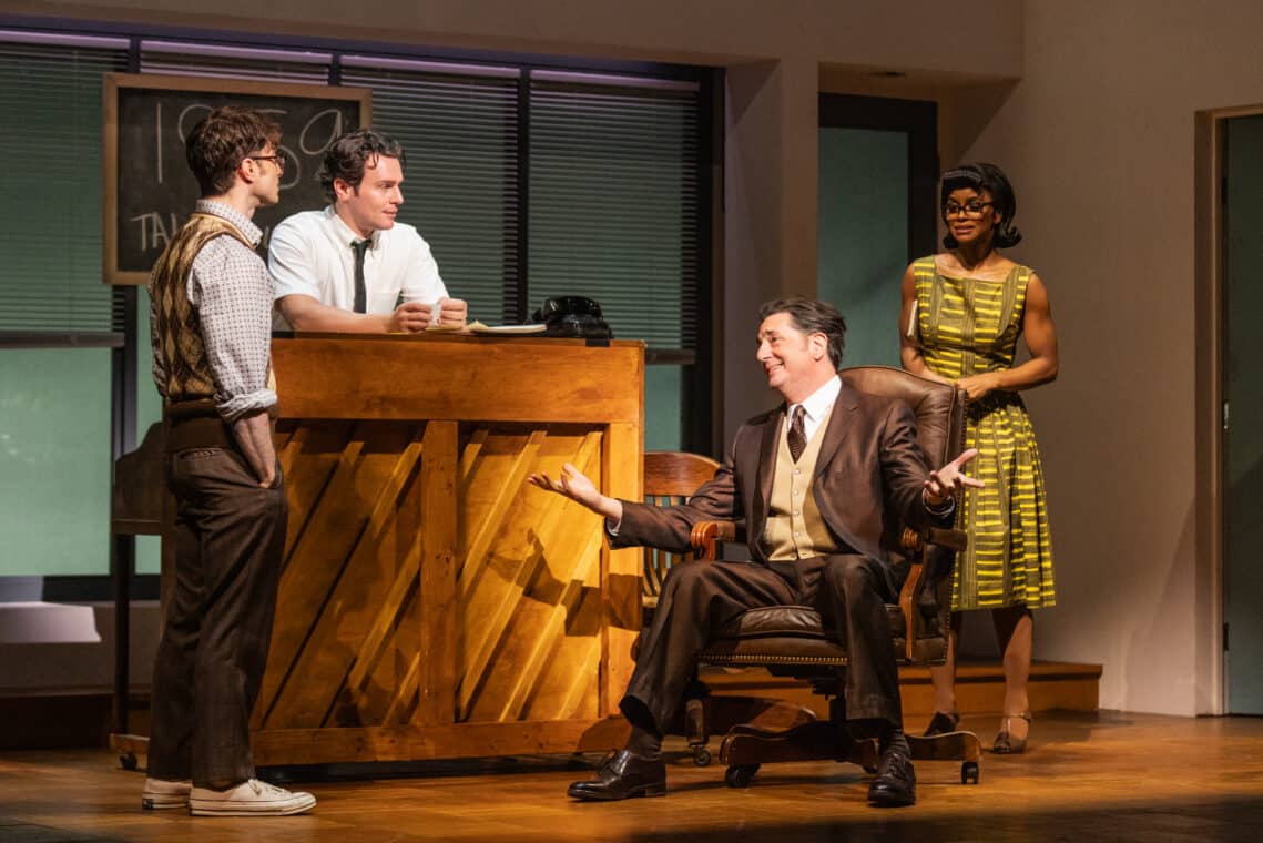 A man in a brown suit is speaking to two young man in show titled Merrily We Roll Along