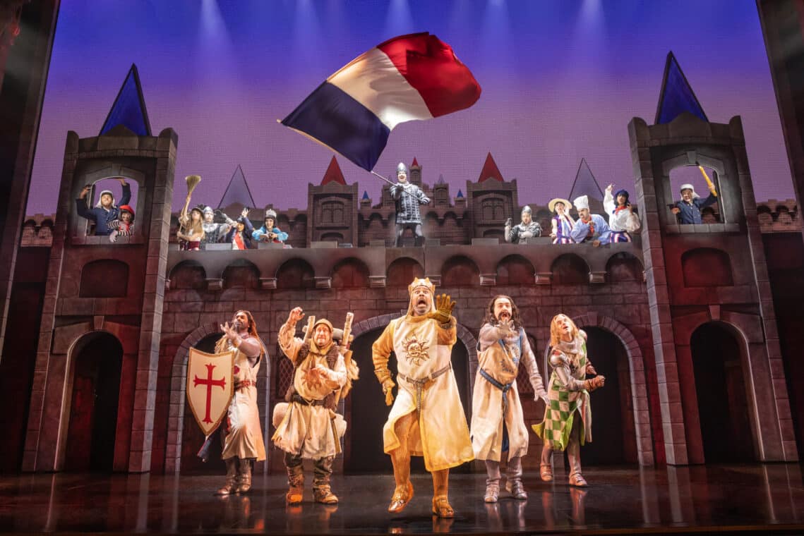 The cast of the Broadway musical Spamalot is standing in front of a palace, waving a French flag