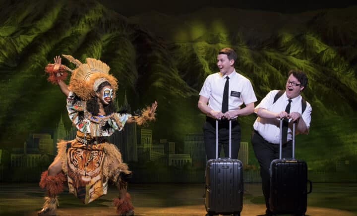 Two men in white shirts laugh as a performer in vibrant costume dances near them on stage in The Book of Mormon