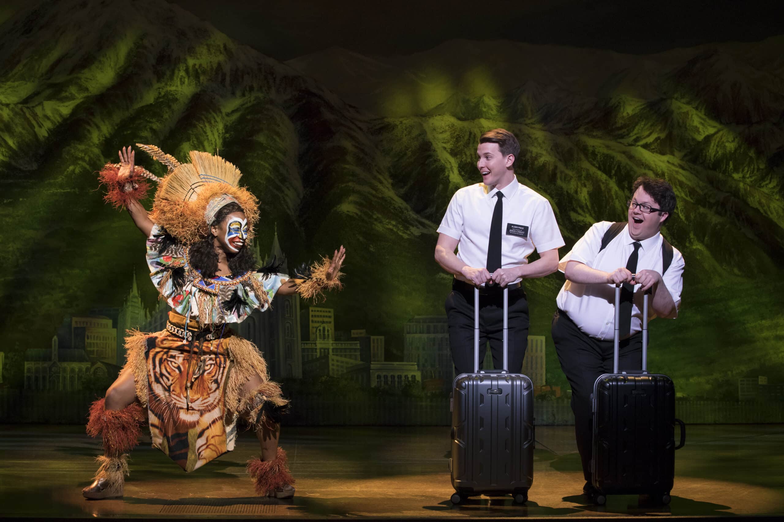 Two men in white shirts laugh as a performer in vibrant costume dances near them on stage in The Book of Mormon