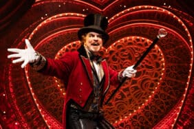 A performer in a red velvet coat and top hat sings with a cane on stage in Moulin Rouge! The Musical