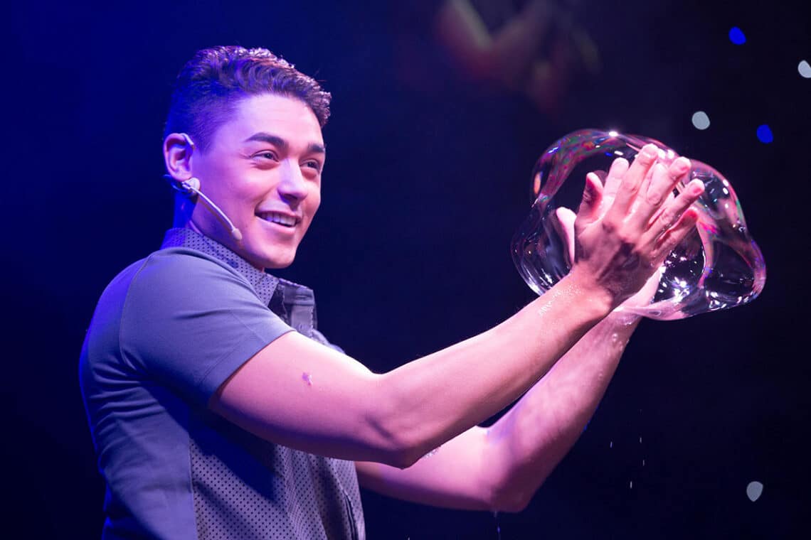 A performer with a headset microphone manipulates a large bubble on stage in The Gazillion Bubble Show