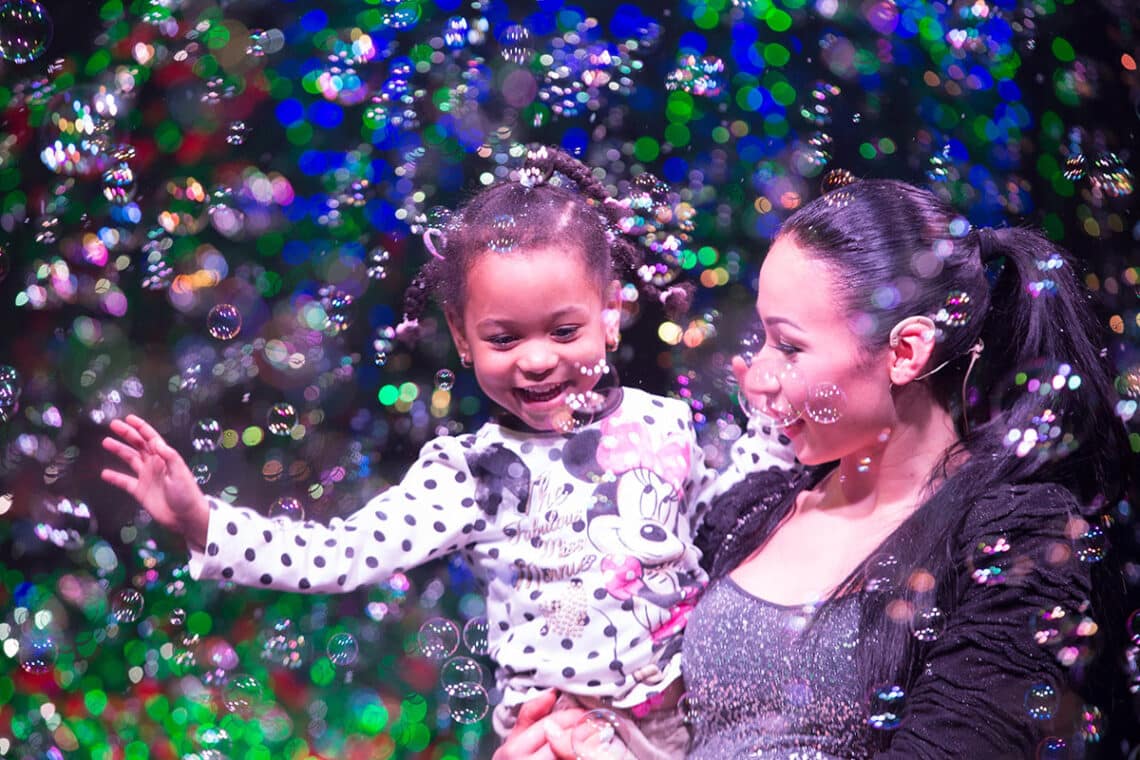 A woman and a young girl smile while surrounded by bubbles on stage in The Gazillion Bubble Show