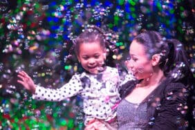 A woman and a young girl smile while surrounded by bubbles on stage in The Gazillion Bubble Show