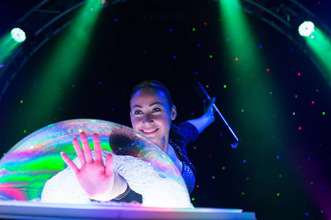 A performer smiles as she creates a large, colorful bubble on stage in The Gazillion Bubble Show