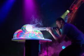 A performer blows into a tube, creating colorful, smoke-filled bubbles on stage in The Gazillion Bubble Show