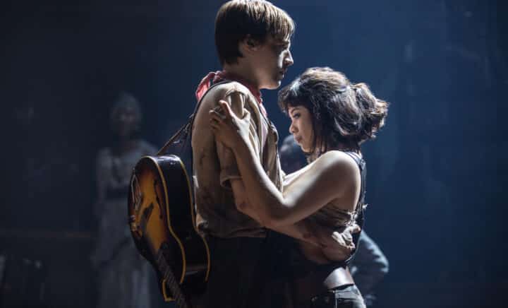 Two actors embrace intimately on stage at Hadestown, with one holding a guitar under dim lighting