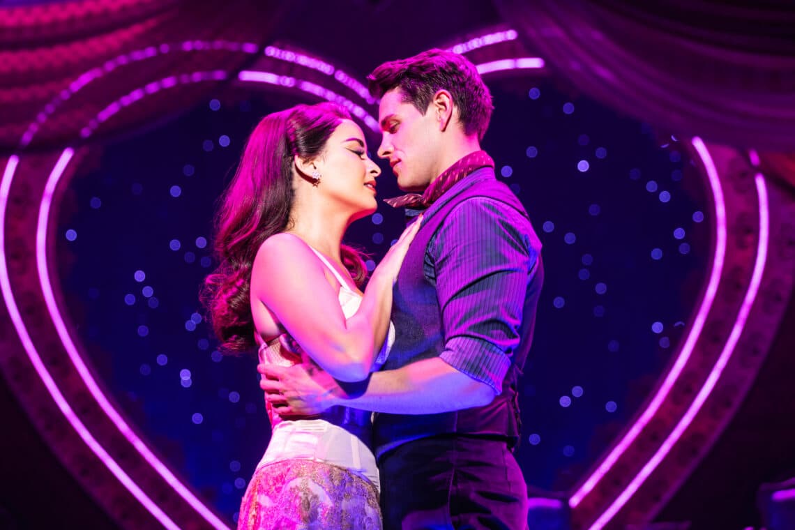 Characters from Moulin Rouge! The Musical share a romantic moment on stage, with vibrant pink and purple lighting