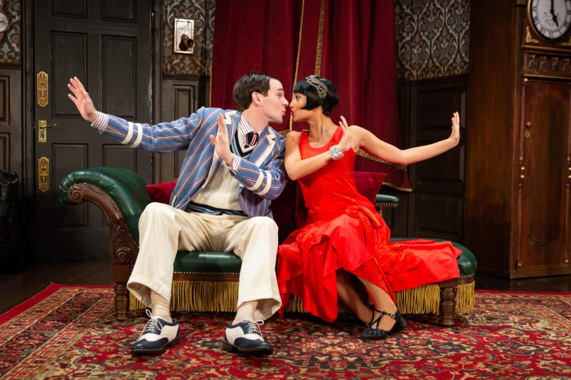 Scene from The Play That Goes Wrong with two actors seated on a couch, humorously posing as if to kiss