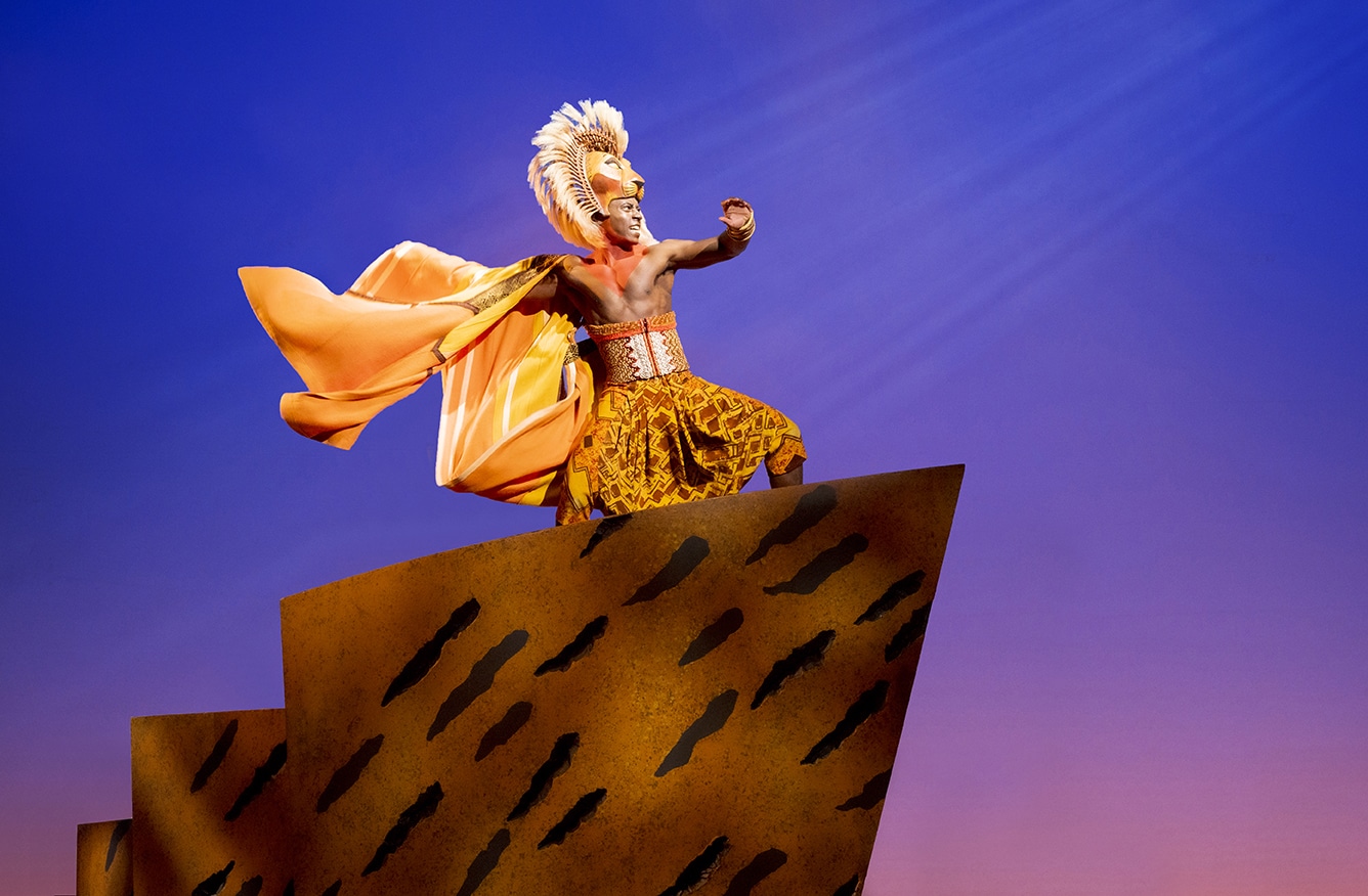 Scene from The Lion King musical with a performer in costume standing proudly on Pride Rock against a gradient sky