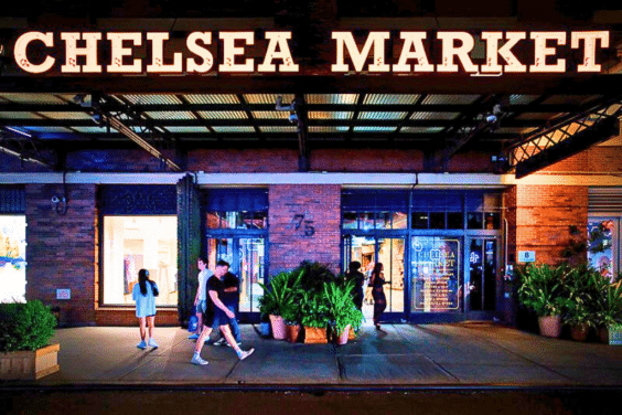 Chelsea Market in NYC at night, featuring bright lights, storefronts, and pedestrians walking along the sidewalk