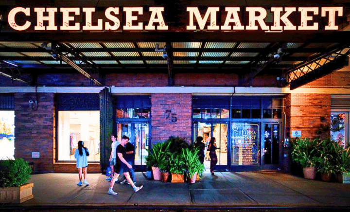 Chelsea Market in NYC at night, featuring bright lights, storefronts, and pedestrians walking along the sidewalk