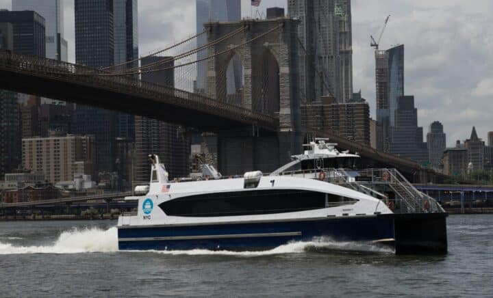 East River ferry with Brooklyn Bridge and Manhattan buildings in the background on a cloudy day