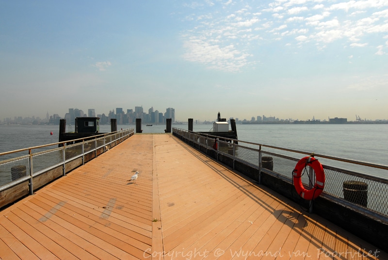 NYC skyline seen from Ellis Island, looking down a wooden pier towards the city on a clear day