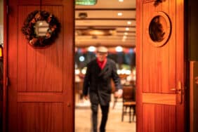 Man with a red scarf and hat walks out a wooden swing door on a cruise ship with a Christmas wreath