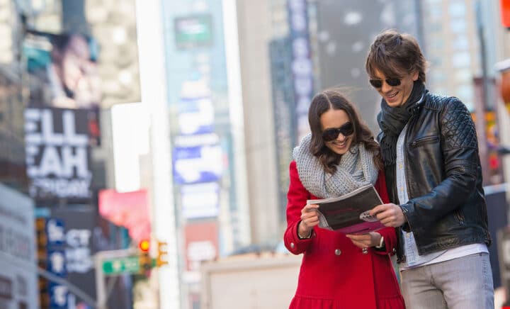 Young tourist couple exploring Times Square, New York City, holding a newspaper and smiling. Why Visit New York City - discover 6 Reasons to Visit NYC including iconic attractions, vibrant culture, and endless entertainment.