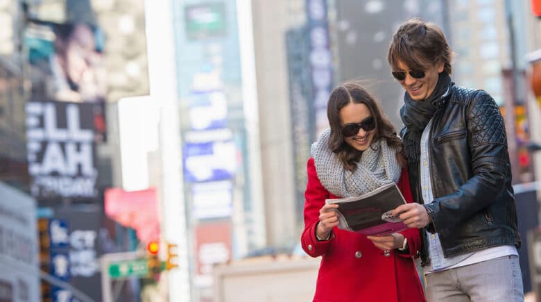 Young tourist couple exploring Times Square, New York City, holding a newspaper and smiling. Why Visit New York City - discover 6 Reasons to Visit NYC including iconic attractions, vibrant culture, and endless entertainment.