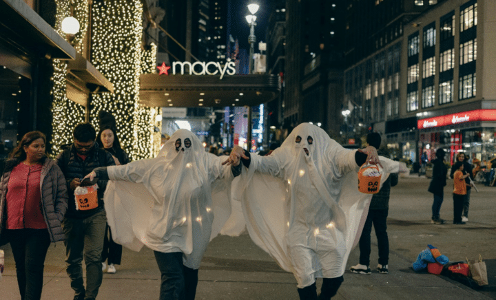 People dressed as ghosts in NYC holding jack-o-lantern buckets while others walk on the pavement