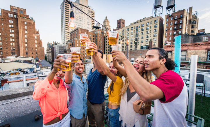 A group of friends celebrating with beer glasses raised in New York City, with the iconic skyline in the background. The party atmosphere is festive, with people laughing, dancing, and clinking glasses. The image suggests a fun and exciting weekend in NYC, with events like Oktoberfest and Blocktoberfest taking place.