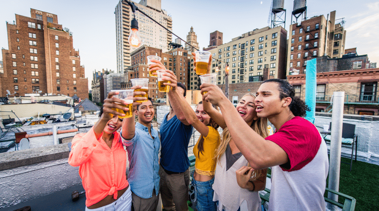 A group of friends celebrating with beer glasses raised in New York City, with the iconic skyline in the background. The party atmosphere is festive, with people laughing, dancing, and clinking glasses. The image suggests a fun and exciting weekend in NYC, with events like Oktoberfest and Blocktoberfest taking place.