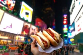 A close-up of a hand holding two hot dogs with mustard and ketchup, with the vibrant lights of Times Square in the background. Mini Wagyu Hot Dogs are jus one of the delicious offerings you will find at Taste of Times Square 2024.