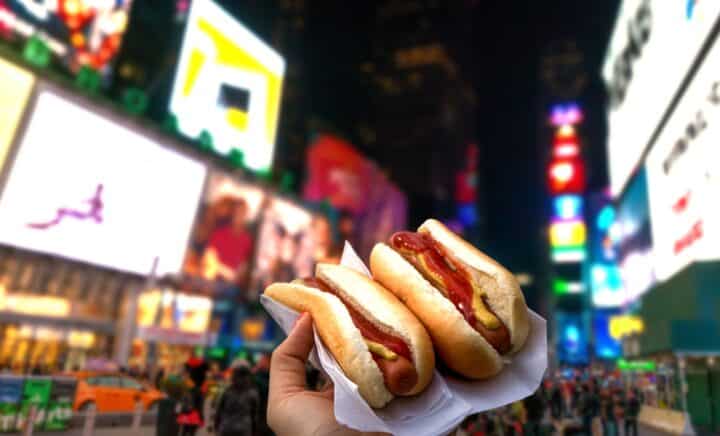 A close-up of a hand holding two hot dogs with mustard and ketchup, with the vibrant lights of Times Square in the background. Mini Wagyu Hot Dogs are jus one of the delicious offerings you will find at Taste of Times Square 2024.