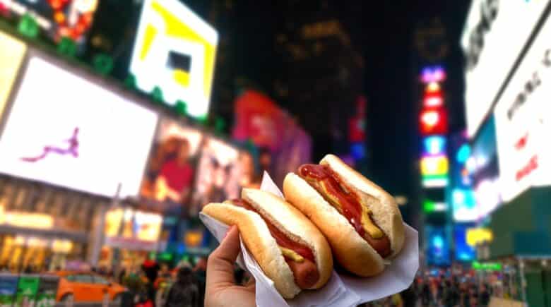 A close-up of a hand holding two hot dogs with mustard and ketchup, with the vibrant lights of Times Square in the background. Mini Wagyu Hot Dogs are jus one of the delicious offerings you will find at Taste of Times Square 2024.