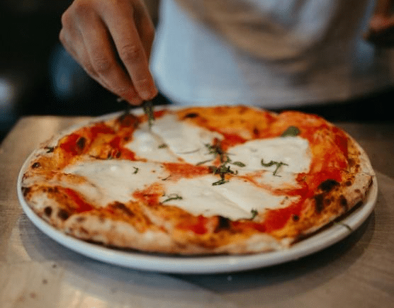 A bustling New York City pizzeria with chefs preparing pizza and a variety of fresh ingredients on display