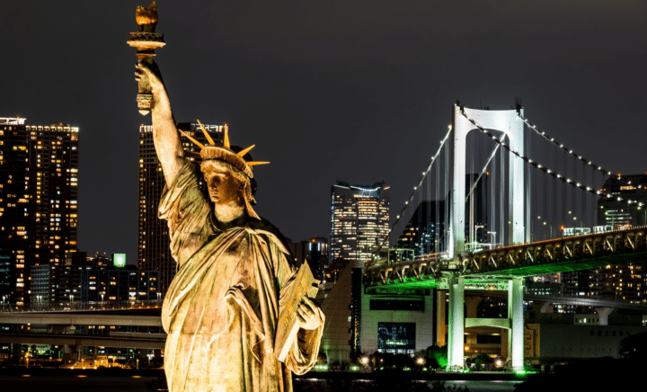 The Statue of Liberty illuminated against the night sky with the skyline and bridge in the background showcasing the views of a NYC dinner cruise