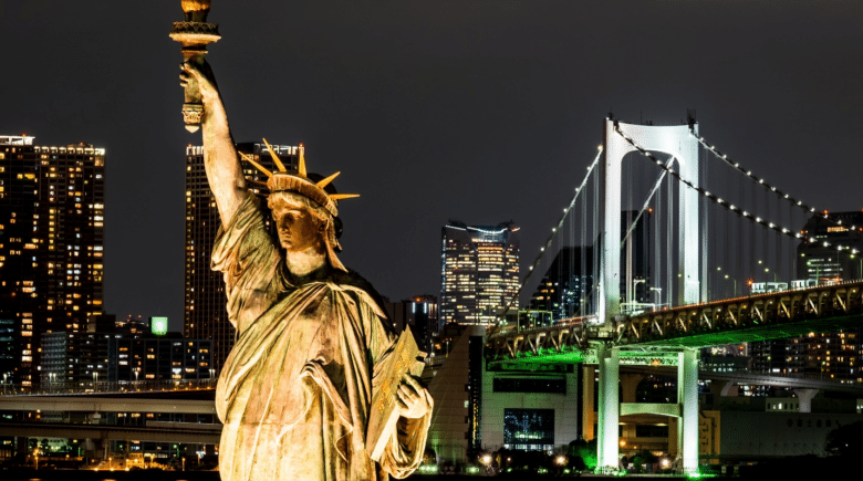 The Statue of Liberty illuminated against the night sky with the skyline and bridge in the background showcasing the views of a NYC dinner cruise