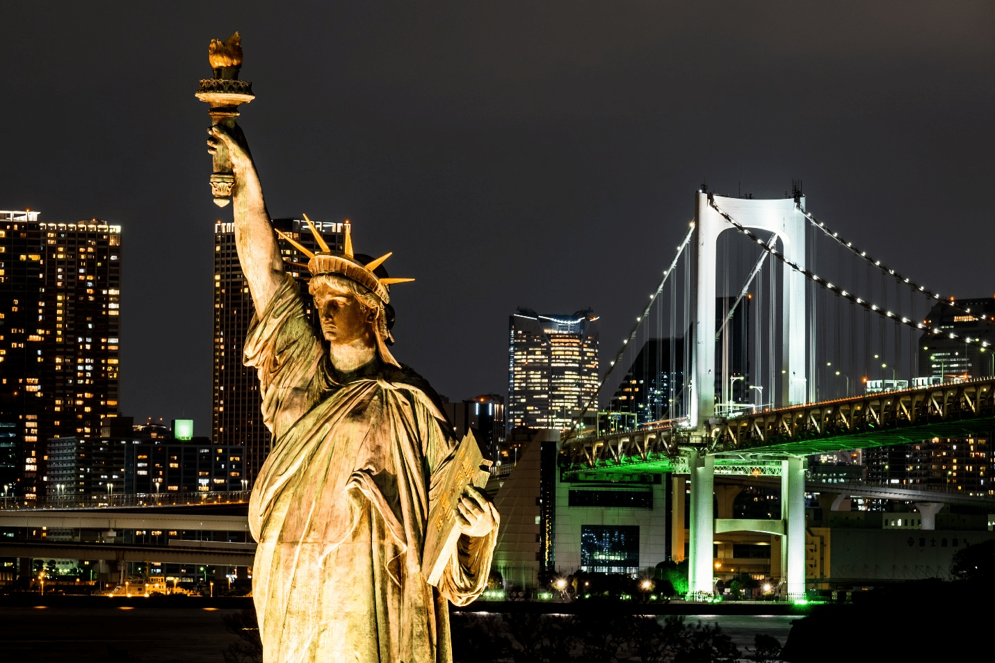 The Statue of Liberty illuminated against the night sky with the skyline and bridge in the background showcasing the views of a NYC dinner cruise
