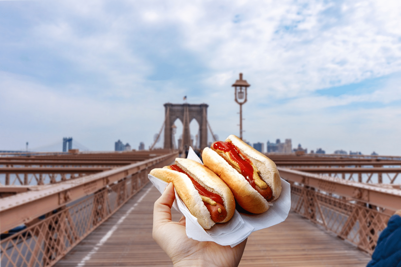 Two hotdogs on a plate with the Brooklyn Bridge in the background.