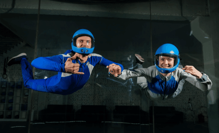 Two smiling indoor skydivers in helmets and suits flying together in a wind tunnel.